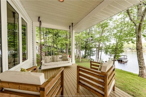 Covered porch with wooden furniture, hanging swing, and view of lake through trees