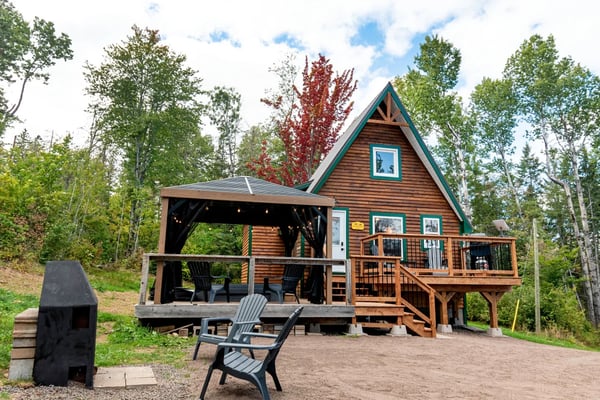 A-frame cabin with brown wood siding and teal trim surrounded by forest, featuring a covered porch, deck, and fire pit with Adirondack chairs.