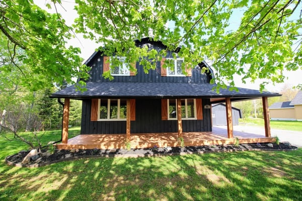 Modern black cabin with wooden porch and shutters, surrounded by green trees and lawn.